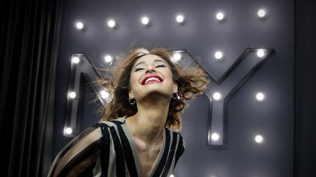 Beautiful model posing for the camera and smiling broadly in the studio on a gray background.  Wind blows the hair of a pretty young girl with red lipstick against the backdrop of many burning light