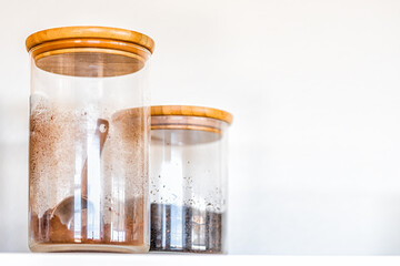 Closeup of two kitchen closed spice glass jars with wooden lids for coffee, cocoa and cinnamon with measuring spoon inside on shelf against white background