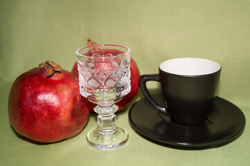 A small crystal glass, a black coffee Cup and saucer, and red ripe pomegranates on a gray-green background.