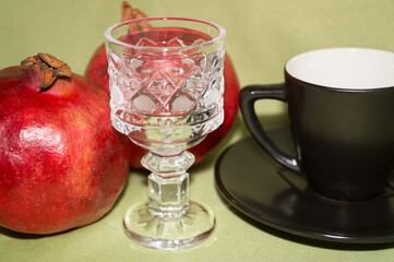 A small crystal glass, a black coffee Cup and saucer, and red ripe pomegranates on a gray-green background.