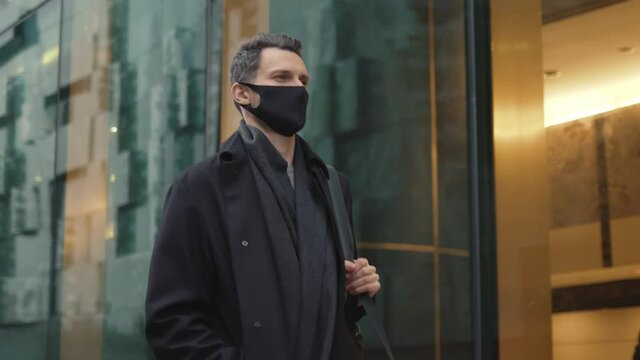Man in black coat and black protective mask walking along the street, slow motion. City street with glass windows of office building on background