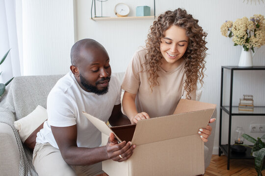 Multinational Couple Looks Forward To Opening A Parcel Ordered Via The Internet. Husband And Pregnant Wife, Sitting On The Sofa, Take Out The Box Capacity. Family, Pregnancy And Relationships Concept.