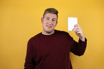 Young blonde man wearing a casual red sweater over yellow background pointing blank notebook in her hand