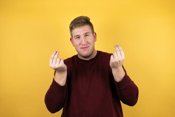 Young blonde man wearing a casual red sweater over yellow background doing money gesture with...