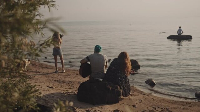 A group of hipster people spend holidays together at sea. The guy playing guitar to his friends at the backdrop of sunset. A girl photographs a guy in a river. isolation together, vacation concept.