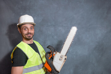 A funny man holds a chainsaw in his hands and looks at the camera. A worker in a signal vest and a helmet.