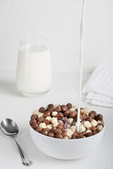 Bowl full of ready-to-eat cold cereals in shape of balls served with spoon and glass of milk with liquid pouring in food prepared for breakfast on white wooden table in the morning. Vertical