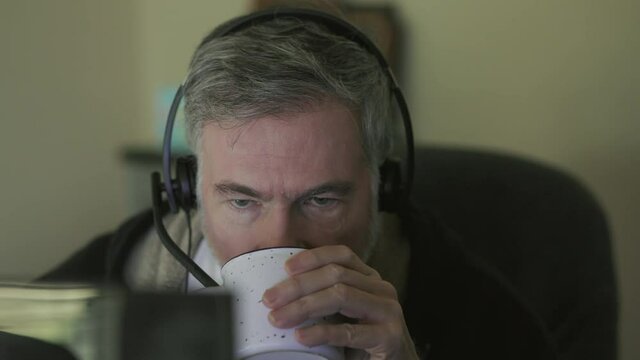 A mature man wearing headphones and drinking morning coffee is engaged with coworkers virtually via his computer which is the new normal of working from home due to the pandemic.