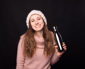 Smiling girl wearing winter clothes is holding a metal thermos over black background.