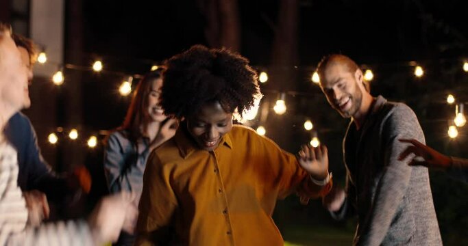 African American young curly woman dancing and jumping among mixed-races friends at night. Late evening party. Pretty female dance and having fun with multi ethnic people.
