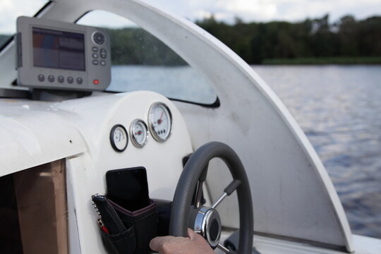 Man's hand on floating motor boat steering wheel, cabin watercraft interior dashboard with control appliances and chart plotter at summer day on water and river shore background, outdoor travel