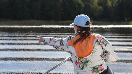 Tourist caucasian young woman in white cap he points to the shore forest from boat bow deck with railing back view, outdoor active recreation on water in Russia