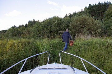Russian mushroomer man  goes for mushrooms with a basket on coast in green grass from boat in forest at summer day, outdoor vacation lifestyle