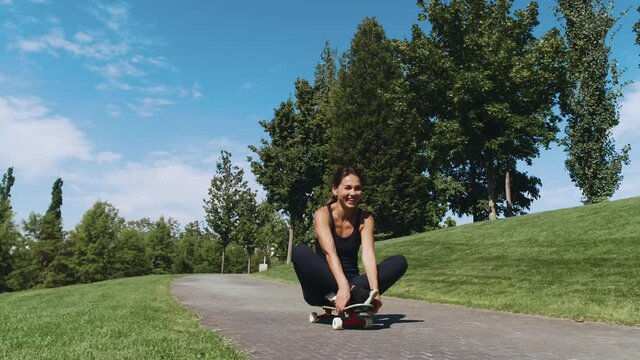 Beautiful fitness girl rides while sitting on a skateboard down.In an open Park