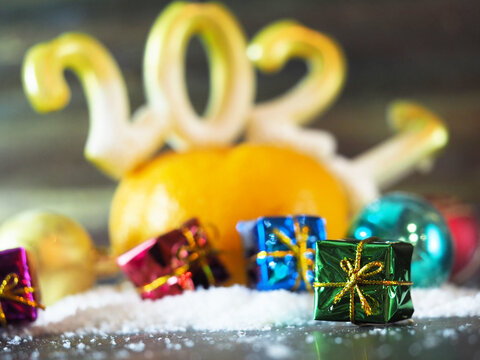 small gifts in multicolored festive shiny paper lie in the snow near a ripe orange tangerine with candles in the form of 2021 numbers. horizontal image with blurred background, soft focus, free space 