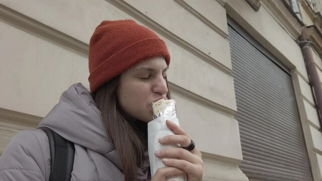 Young woman enjoying eating vegan roll standing by the wall at the street. Street food culture. Go vegan