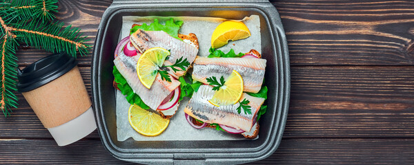 Herring sandwiches with rye bread, parsley and lemon in container for fast food, traditional Danish smorrebrod. Top view, wood background with copy space. Christmas holidays, street snack