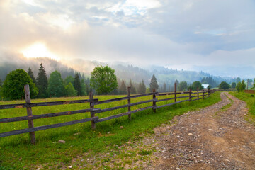Fototapeta na wymiar Road with wooden fence along the meadow, foggy mountains and morning sun. Ukraine, Verkhovyna.