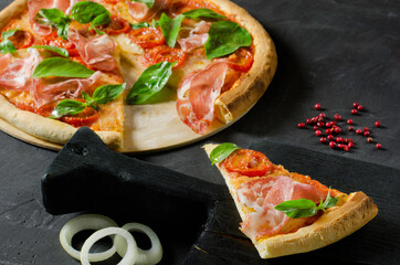 A piece of homemade Italian pizza with prosciutto, tomato, onion, mozzarella, basil and ingridients on a black cutting board