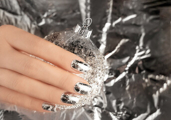 Female hand with trendy silver nail design on foil background holding silver Christmas ball. Festive backdrop for holidays. Holiday silver manicure.