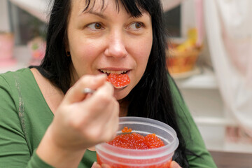The girl eats red caviar with a spoon. A woman is sitting in the kitchen with a spoonful of caviar...