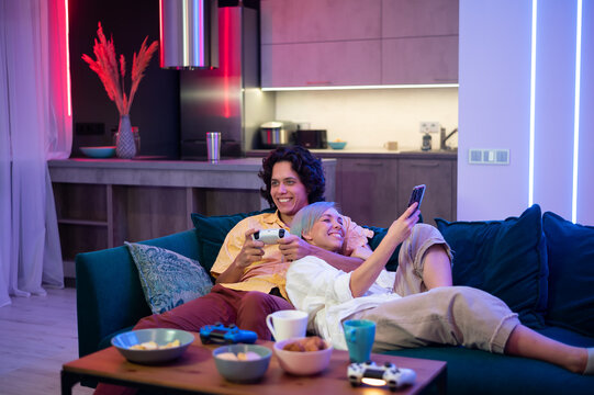 Young woman spending time with her boyfriend at home. She using smart phone while he is playing video games on a gaming console at home.