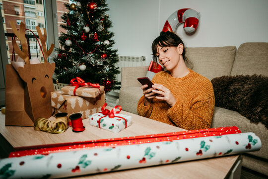 Woman Looking For Information On Her Smartphone While Wrapping Christmas Gifts At Home Accompanied By Her Lovely Dog