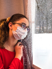 Portrait of a girl during self-isolation due to corona covid-19 virus, teenager girl in a medical mask is sad at the window, quarantine isolation concept. Coronavirus covid-19