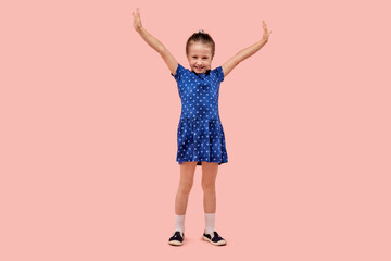 Happy child rejoices in victory hands raised up. The concept of winning. Human emotion. In a blue dress with a small star print. Pink isolated background.