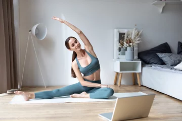 Fototapeten Young sporty fit slim woman coach do practice video online training yoga laptop in living room at home © BestForYou