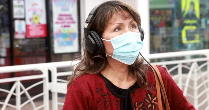 Attractive Woman With Long Dark Hair Wearing A Covid Mask For Coronavirus Epidemic And Headphones In Hand-held Medium Shot. Older Asian Model.