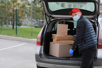 Young delivery man in protective mask, red cap and gloves near the car with boxes and packages, outdoors. Service coronavirus. Online shopping. mock up.