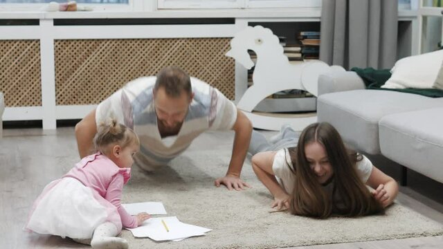 Funny happy family doing push ups together. Mother laughing, father doing fitness, little cute daughter drawing painting on a floor. Happy family funny moments at home while quarantine lockdown. 