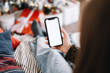 Woman holding a smartphone with a white screen mock up, sitting on the bed at home near Christmas tree.
