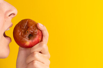 The girl bites a rotten apple with a worm on a yellow background. Expired products, junk food