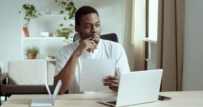Closeup pensive black guy african american businessman mixed race student with laptop wears white t-shirt reads document holds sheet of paper in hands receives letter from bank makes notes in notebook