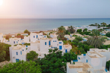 top view of white houses with blue windows and doors, standing on the seashore, among high palm trees. Tunisian city, beautiful white buildings. Rest, resort, travel.