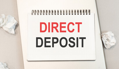 The word direct deposit is written in a notebook that sits on a gray desktop along with a laptop.