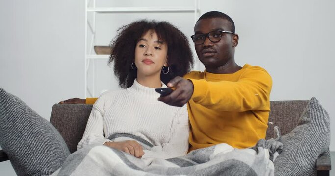 African American family couple ethic man and woman chooses TV channel switches movies programs sits together in embrace on couch sofa in living room at home resting relaxes covered by warm blanket