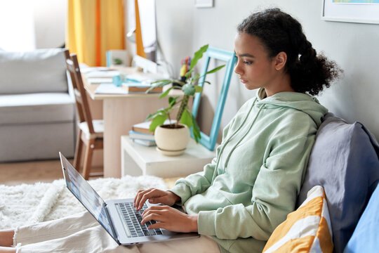 African teen school girl student virtual distance learning online digital class looking at laptop sitting on bed at home. Mixed race gen z teenager remote using computer, typing, elearning in bedroom.