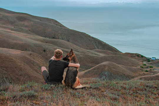 Young man with german shepherd Dog Looking at Sea. Best friends traveling together. Trekking, travel and activity concept.  Recreation and healthy active lifestyle outdoors. Social Distance.