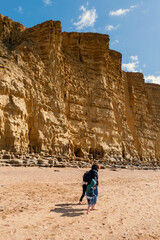 People walking on golden beach underneath towering cliffs on sunny summer day. Jurassic coastline of West Bay in Dorset. UK