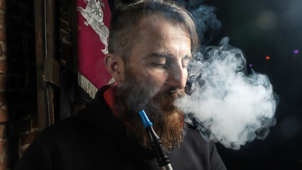 Brutal man with large beard and fashionable haircut smokes an electronic cigarette or hookah on dark background. Steam, vapor, hookah.