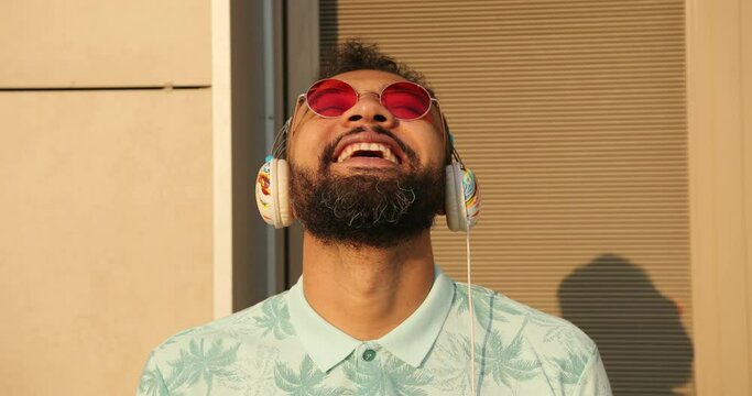 Close-up portrait of the young emotional african man in stylish pink sunglasses listening to music in headphones and lughing outdoor.