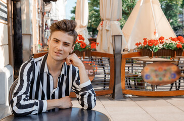 Obraz premium Guy model near the cafe. A young man in stylish clothes is sitting at a table. Cute guy on the summer terrace with flowers.