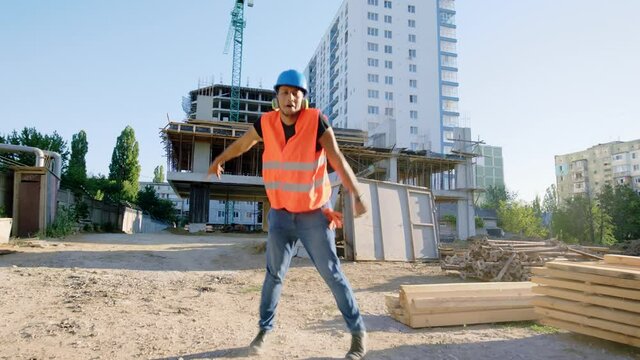 Walking in front of the camera Afro American guy and start dancing at construction site at the lunch time beside his colleagues they all wearing protective uniform