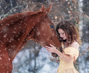 Beautiful curly girl in a yellow dress next to a red horse in winter on a background of snow