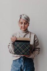 Woman opens Christmas holiday gift box in front of white wall. Green and golden box. Merry Christmas and Happy New Year. Minimalist. Women's hands. Festive gifts. Holiday online shopping.