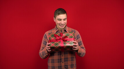 A young man shakes a Christmas present in his hands and rejoices. Shooting in the studio on a red background.