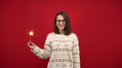 A young woman in a white sweater holds a sparkler in her hand. Shooting in the studio on a red background.